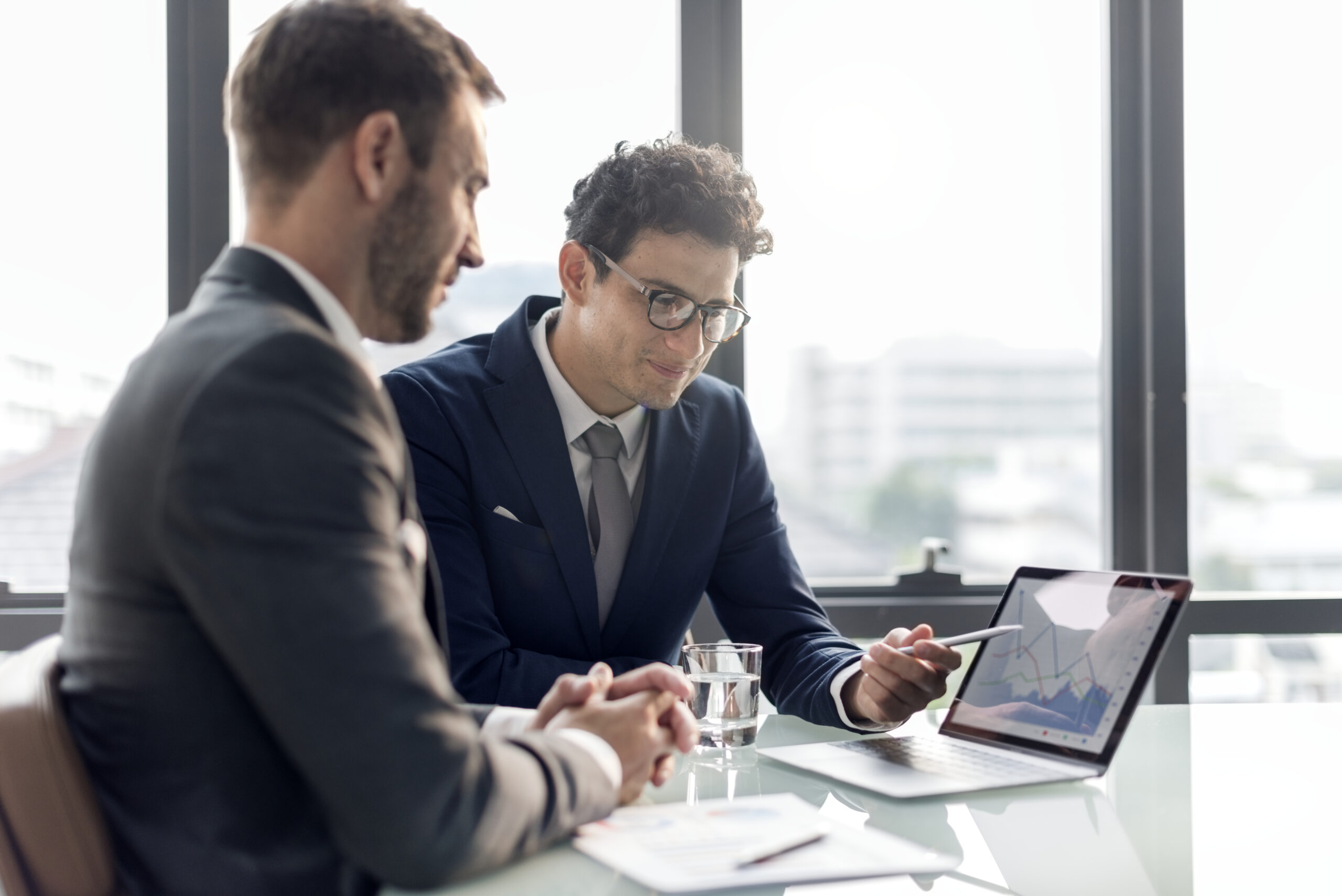 To forretningsmænd i jakkesæt sidder ved et bord i et moderne kontormiljø og diskuterer data vist på en bærbar computer.